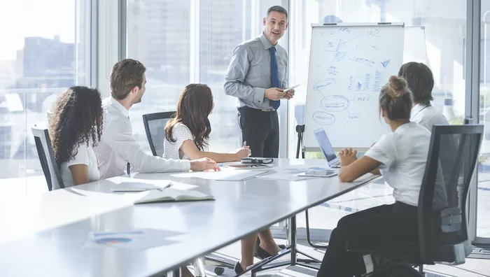 Mature Businessman giving a presentation to his team. He is using a whiteboard with charts and graphs. He is talking. They are in an office boardroom at the table with laptop computers and paperwork. Men and women in the group.